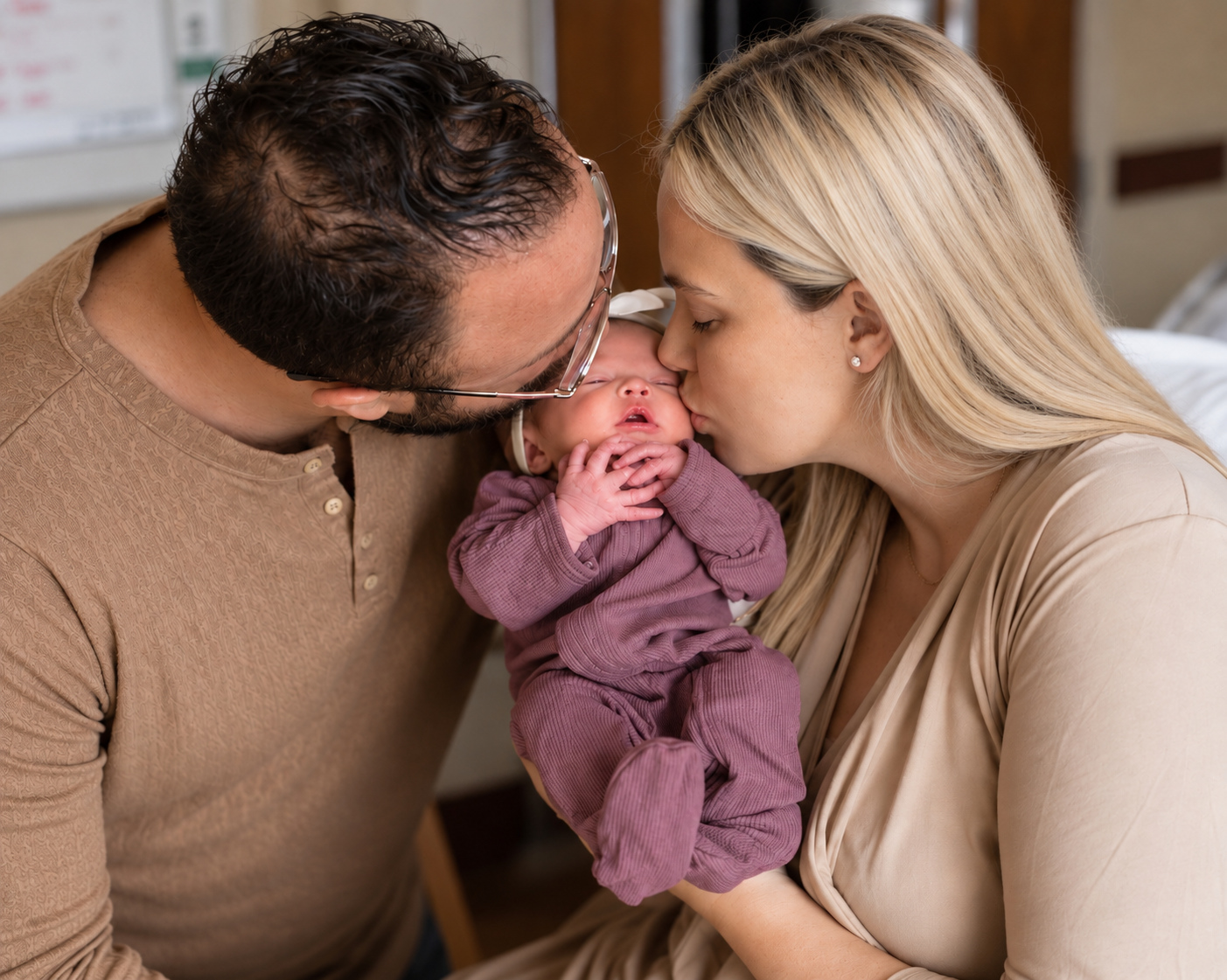 Robbie and Amanda kissing their newborn daughter, who is wearing a plum-colored outfit.